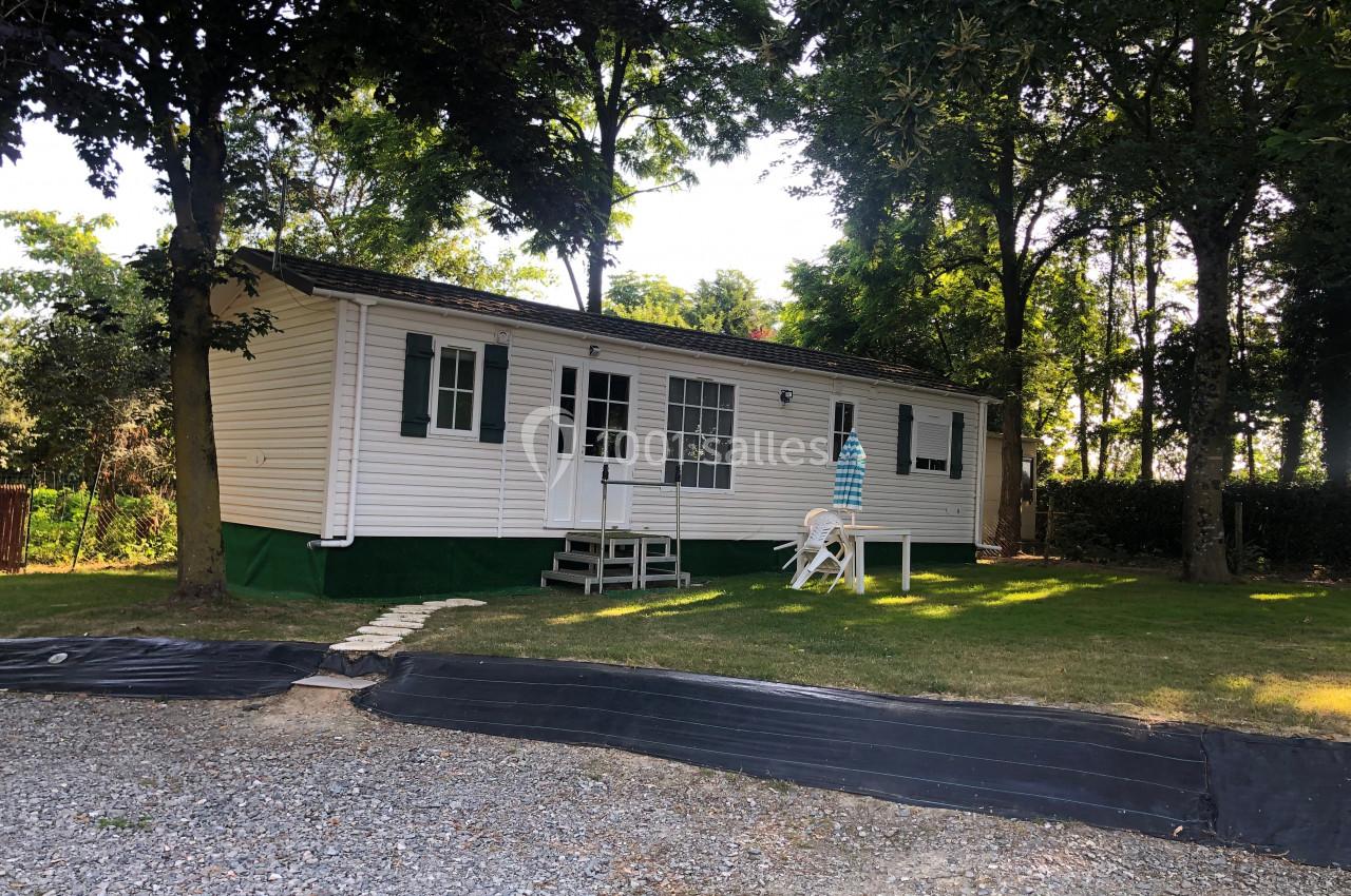 Mobil-home blanc avec terrasse en bois, entouré d'arbres et situé sur une parcelle avec pelouse et allée en gravier.