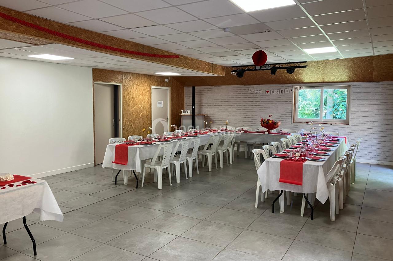 Salle décorée pour un événement, avec tables alignées, nappes blanches et chemins de table rouges, éclairée par la lumière…