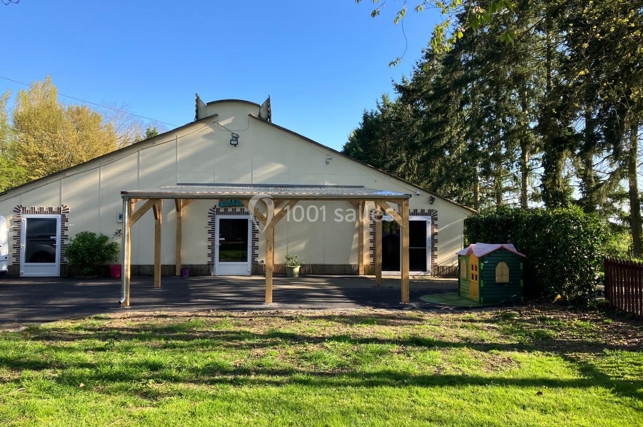 Façade d'un bâtiment clair avec une pergola en bois, entouré d'arbres et d'une pelouse avec une cabane pour enfants.