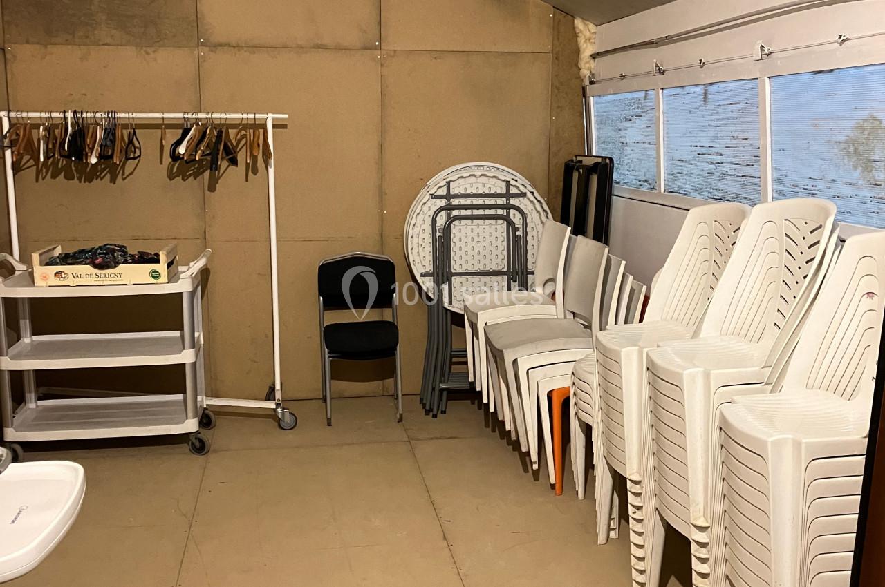 Salle de rangement avec des chaises empilées, une table pliante, un portant à cintres et un chariot à roulettes.