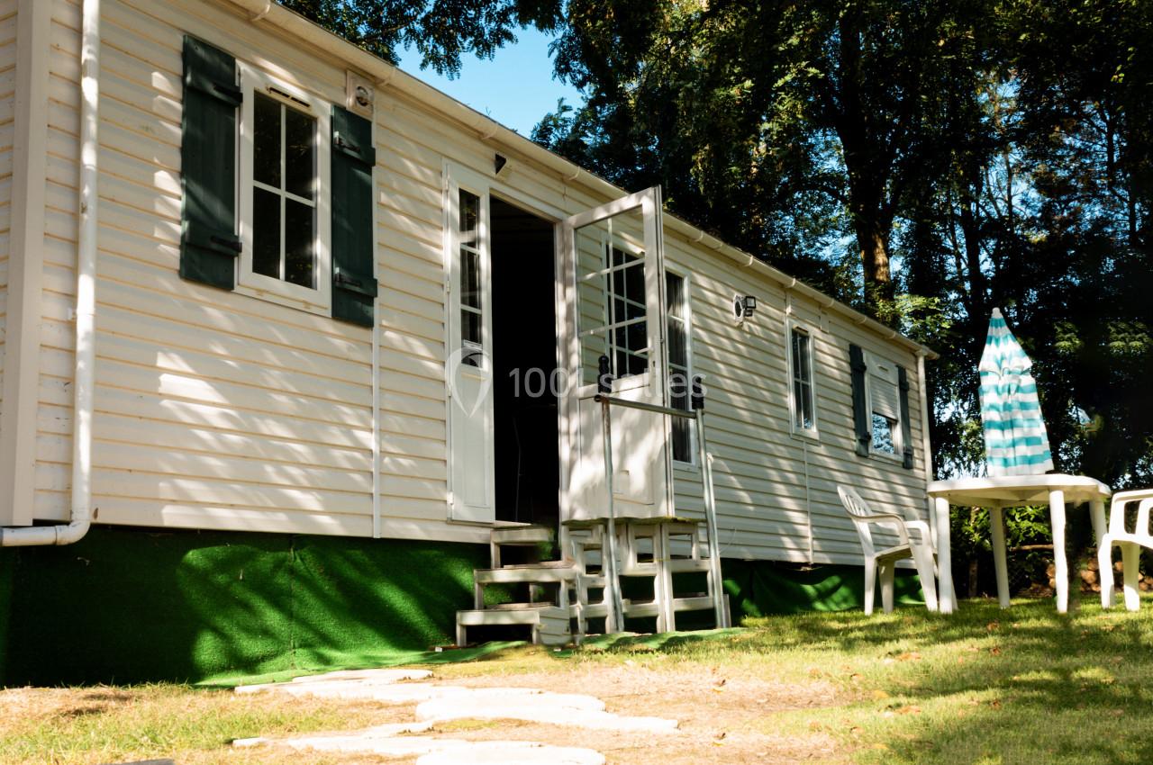 Mobil-home blanc avec porte ouverte, table et chaises en plastique sur une pelouse, entouré d'arbres.