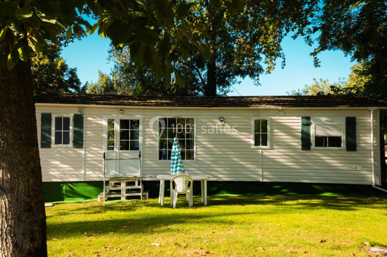 Mobil-home blanc avec volets verts, entouré d'arbres, une table et des chaises en plastique sur la pelouse.