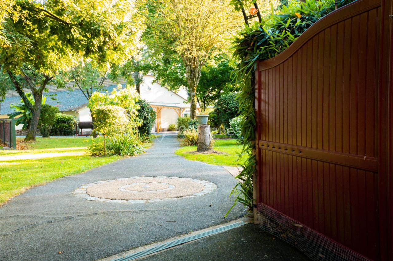 Allée pavée menant à une maison entourée de verdure, vue depuis un portail en bois ouvert.