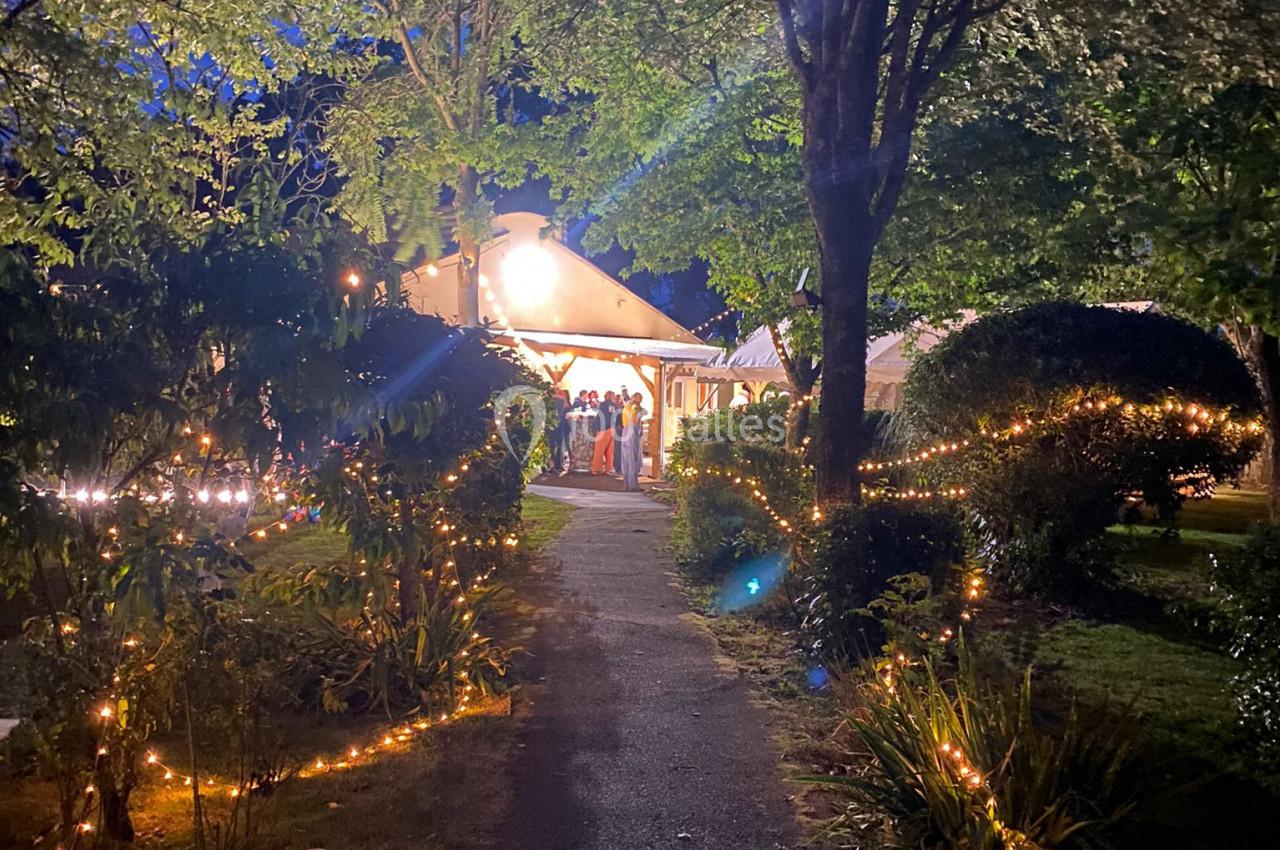 Allée éclairée par des guirlandes lumineuses menant à une maison entourée de végétation, en soirée.