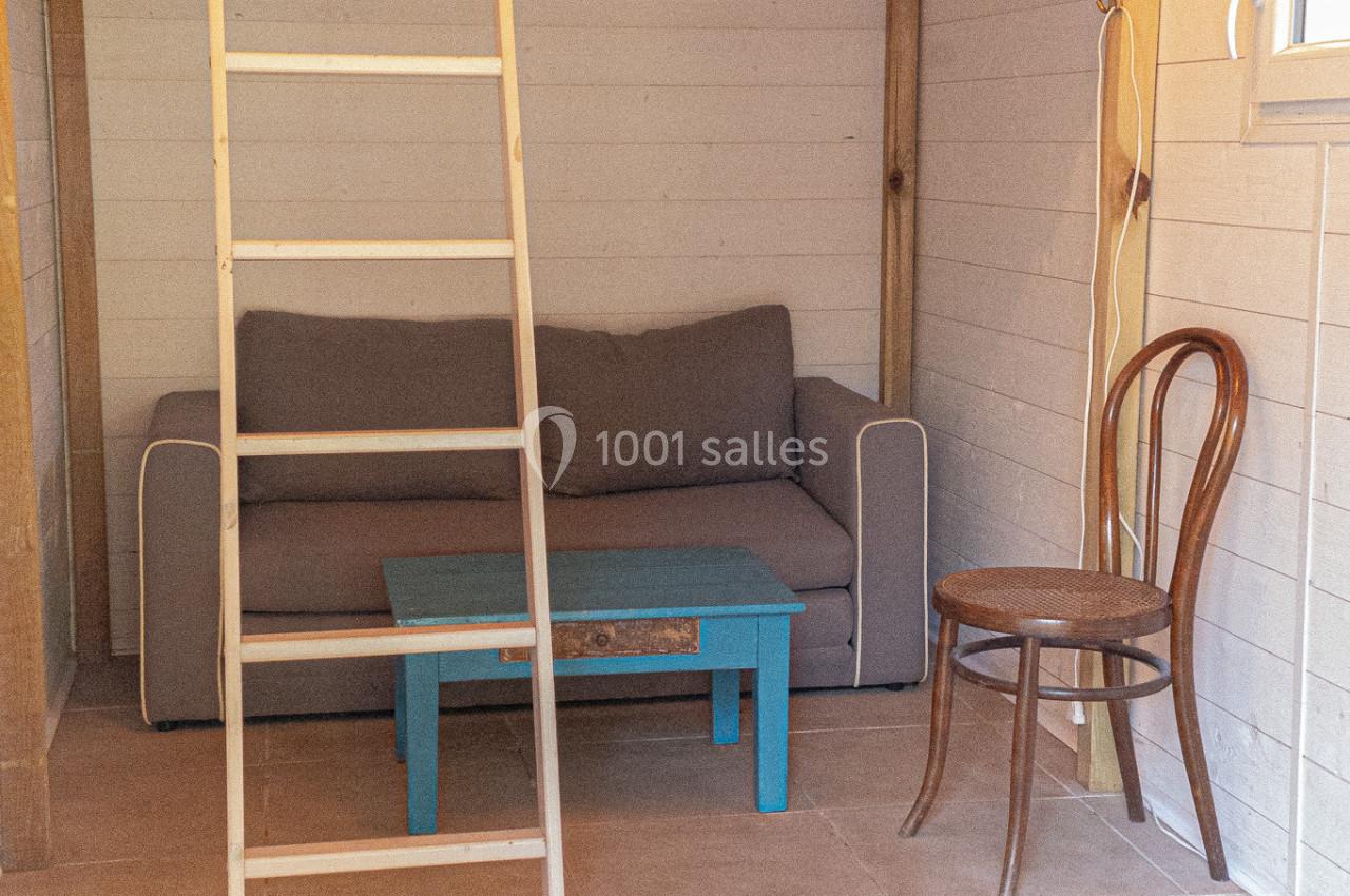 Petit espace aménagé avec un canapé gris, une table basse bleue, une chaise en bois et un lit en mezzanine accessible par…