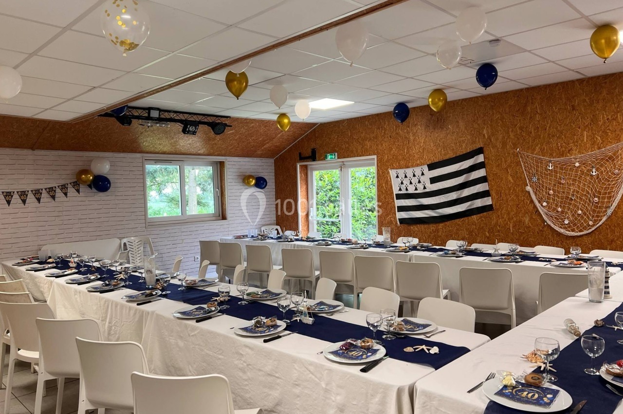 Salle décorée pour un événement avec tables dressées, nappes blanches, décorations bleu et or, et drapeau breton au mur.