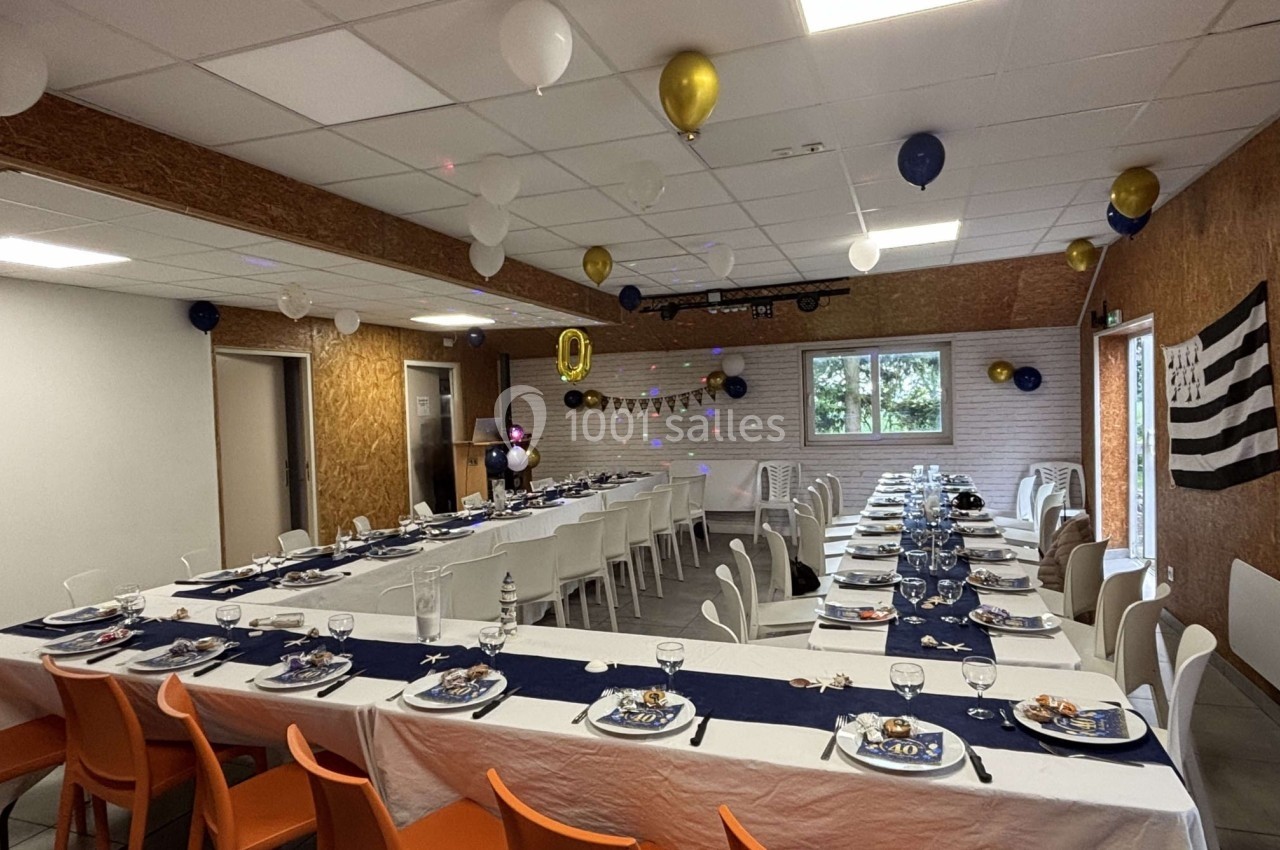 Salle décorée pour une fête avec tables dressées, nappes blanches et bleues, ballons suspendus et drapeau breton au mur.