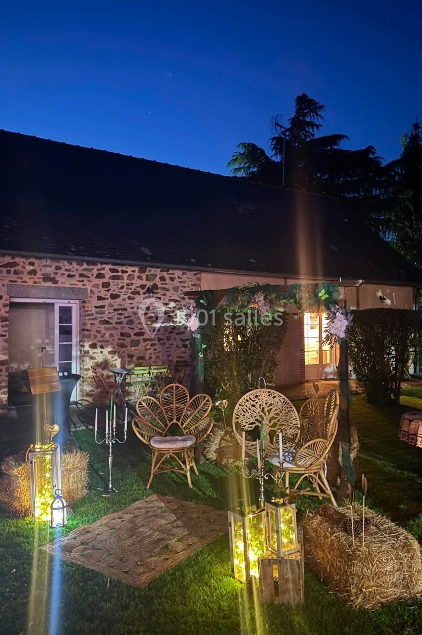 Façade d'une maison en pierre éclairée la nuit, avec des fauteuils en rotin et des lanternes décoratives sur la pelouse.