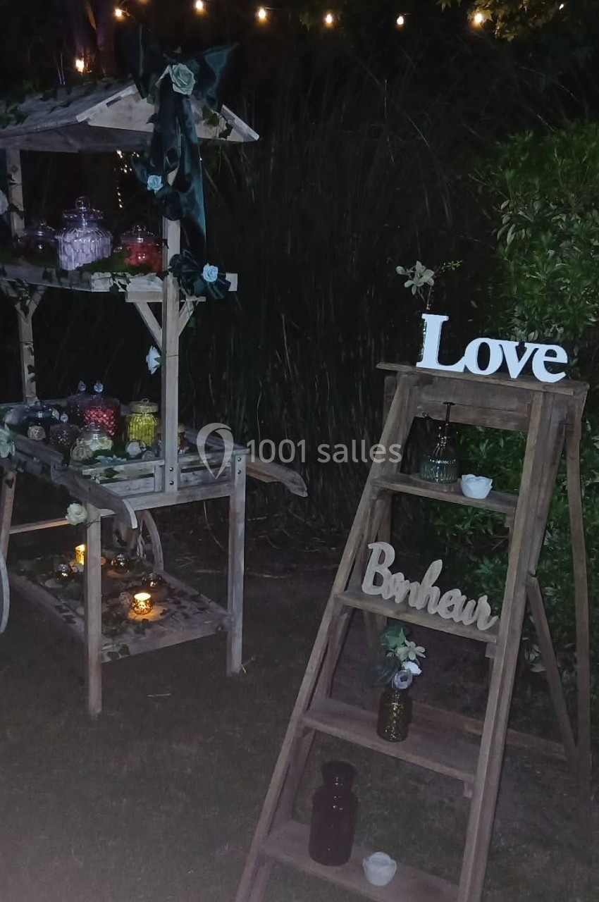 Décoration nocturne avec une échelle en bois portant les mots ’Love’ et ’Bonheur’, et un stand orné de lumières et fleurs.