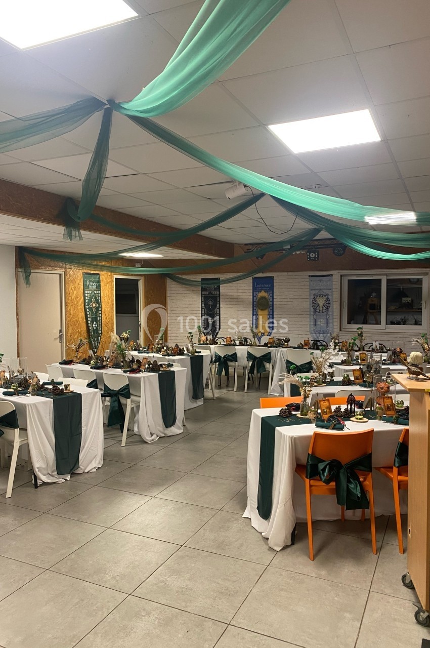 Salle décorée pour un événement, avec des tables dressées, nappes blanches et vertes, et guirlandes suspendues.