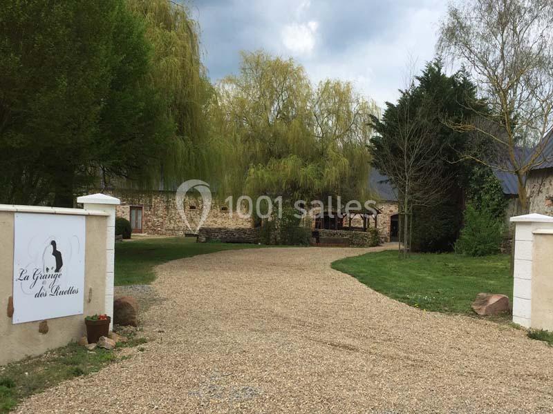 Entrée d'une propriété avec allée en gravier, pelouse, arbres et bâtiments en pierre sous un ciel nuageux.