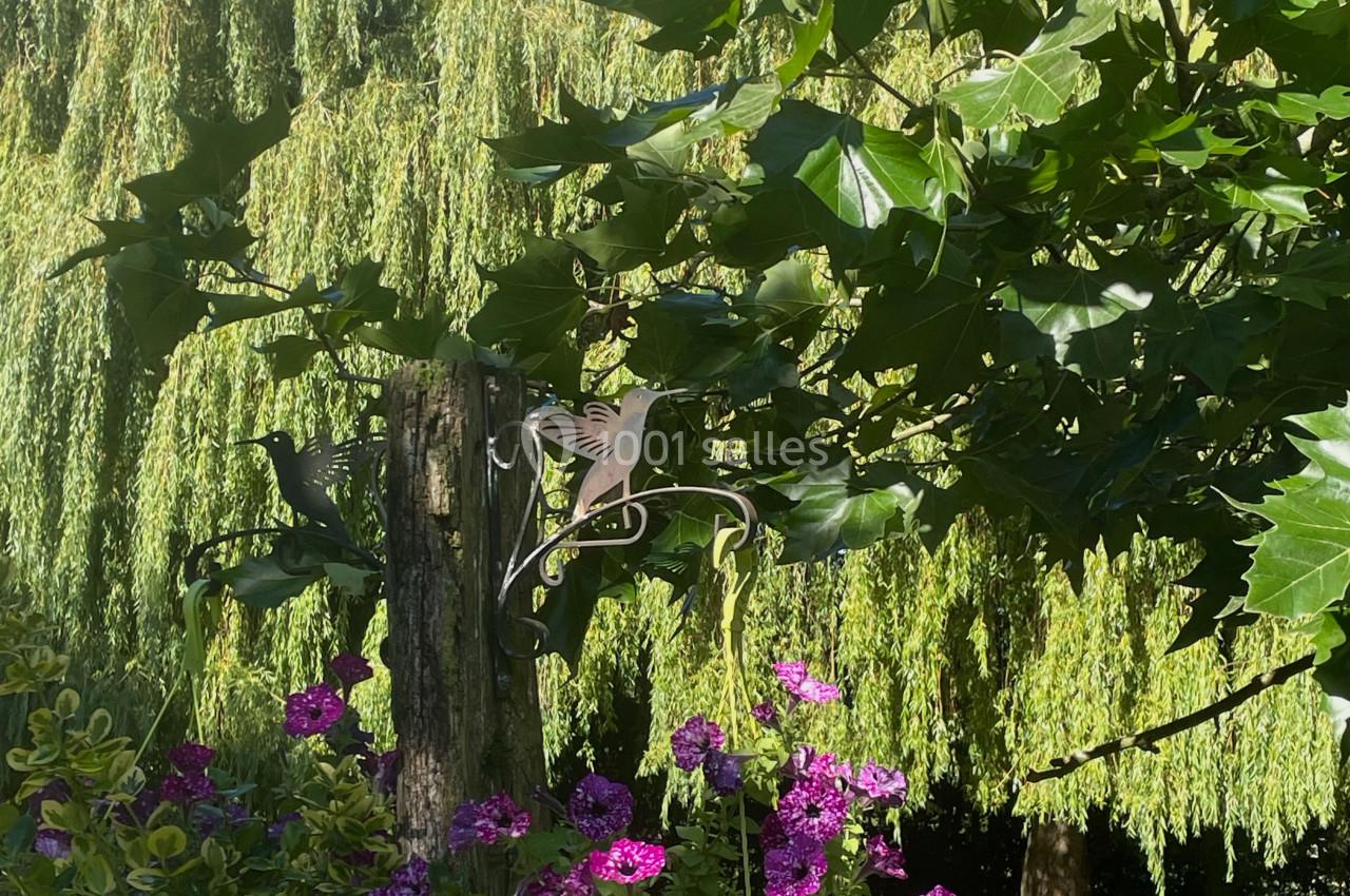 Pot vert avec des fleurs violettes suspendu à un support en métal, devant des arbres et un saule pleureur.