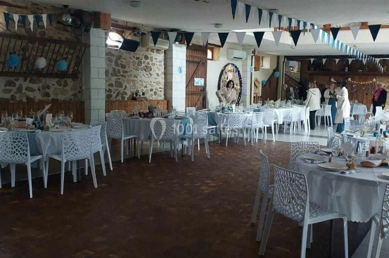 Salle décorée pour une réception avec des tables rondes dressées, des chaises blanches et des guirlandes suspendues.