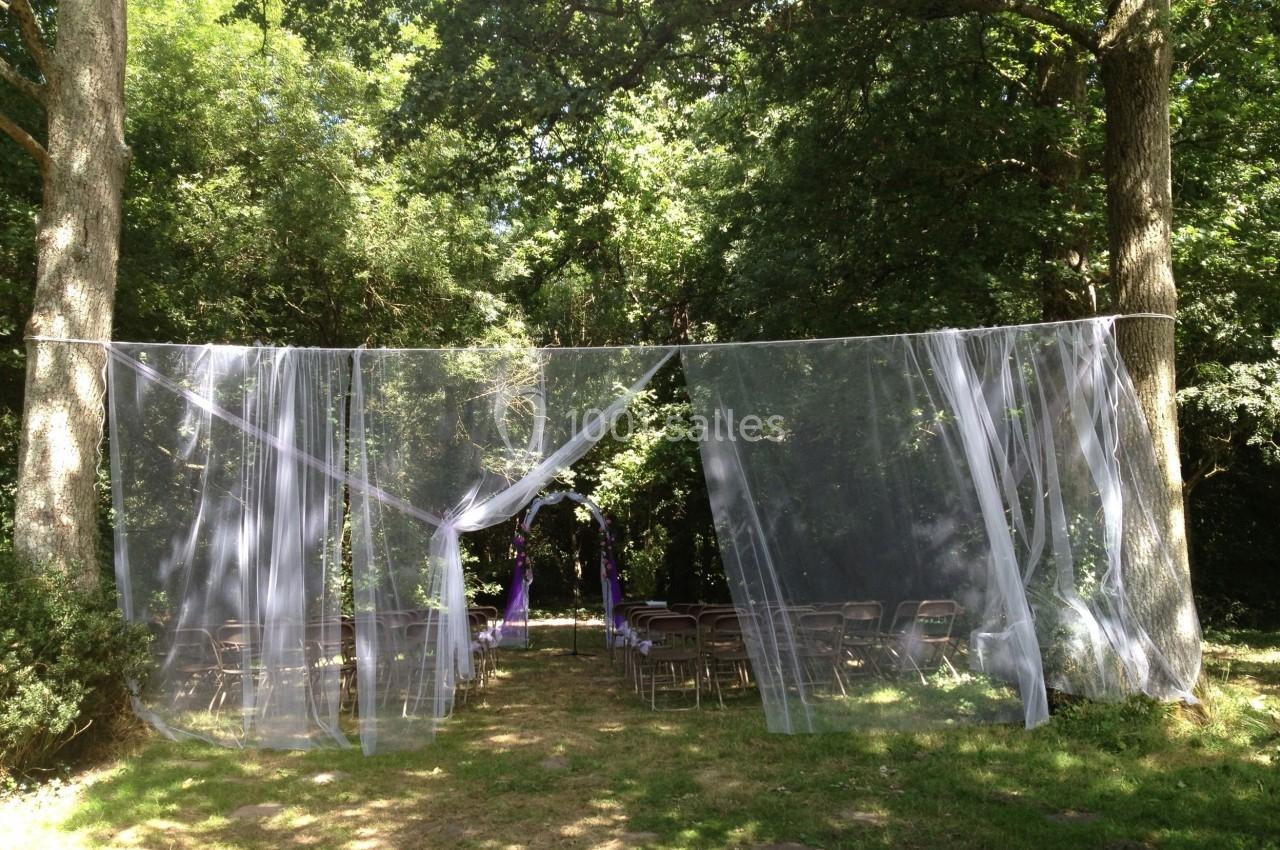 Chaises disposées en extérieur sous des arbres, avec un voile blanc suspendu formant une arche pour une cérémonie.