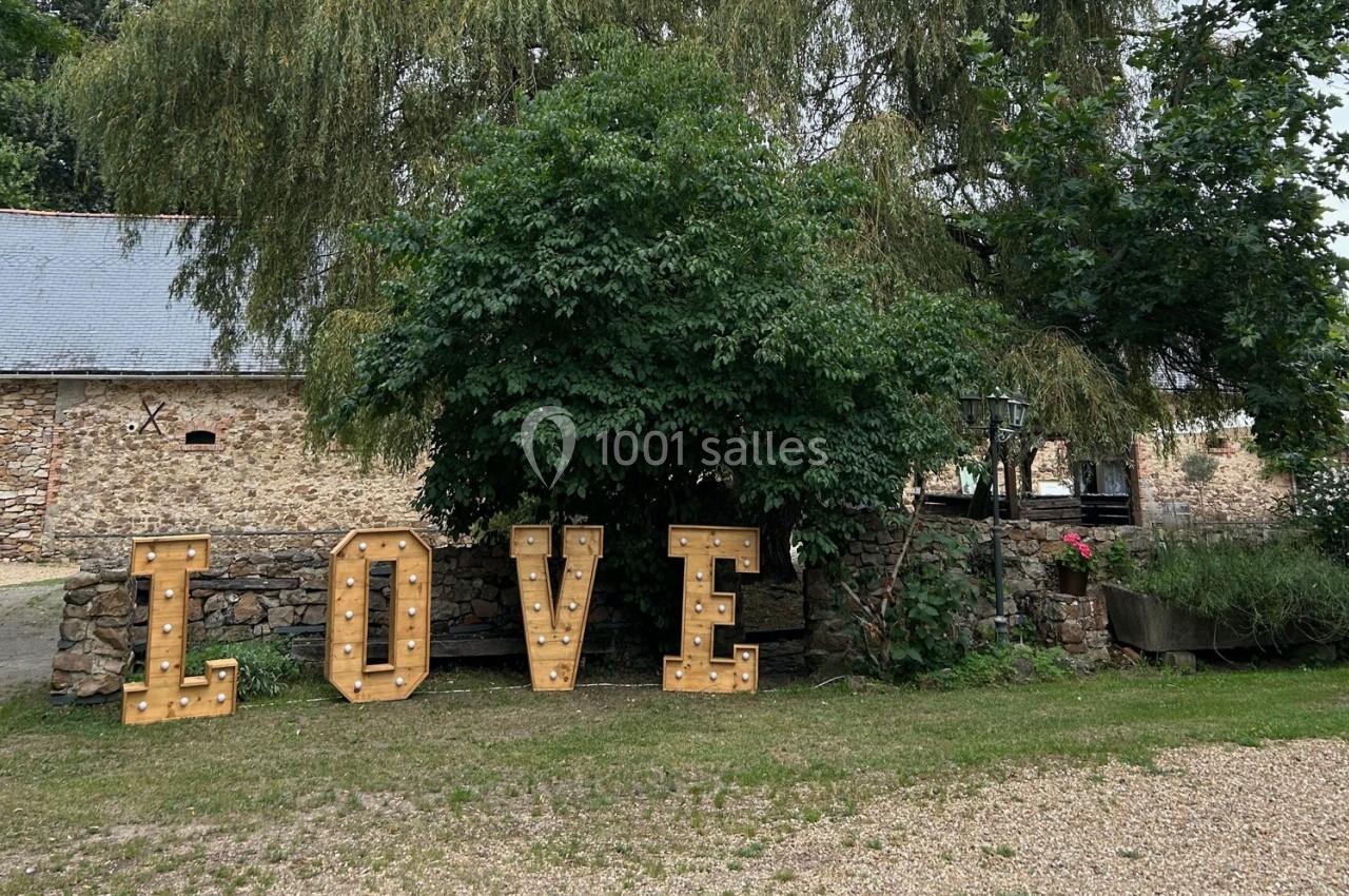 Lettres lumineuses formant le mot ’LOVE’ devant un mur en pierre et des arbres dans un cadre champêtre.