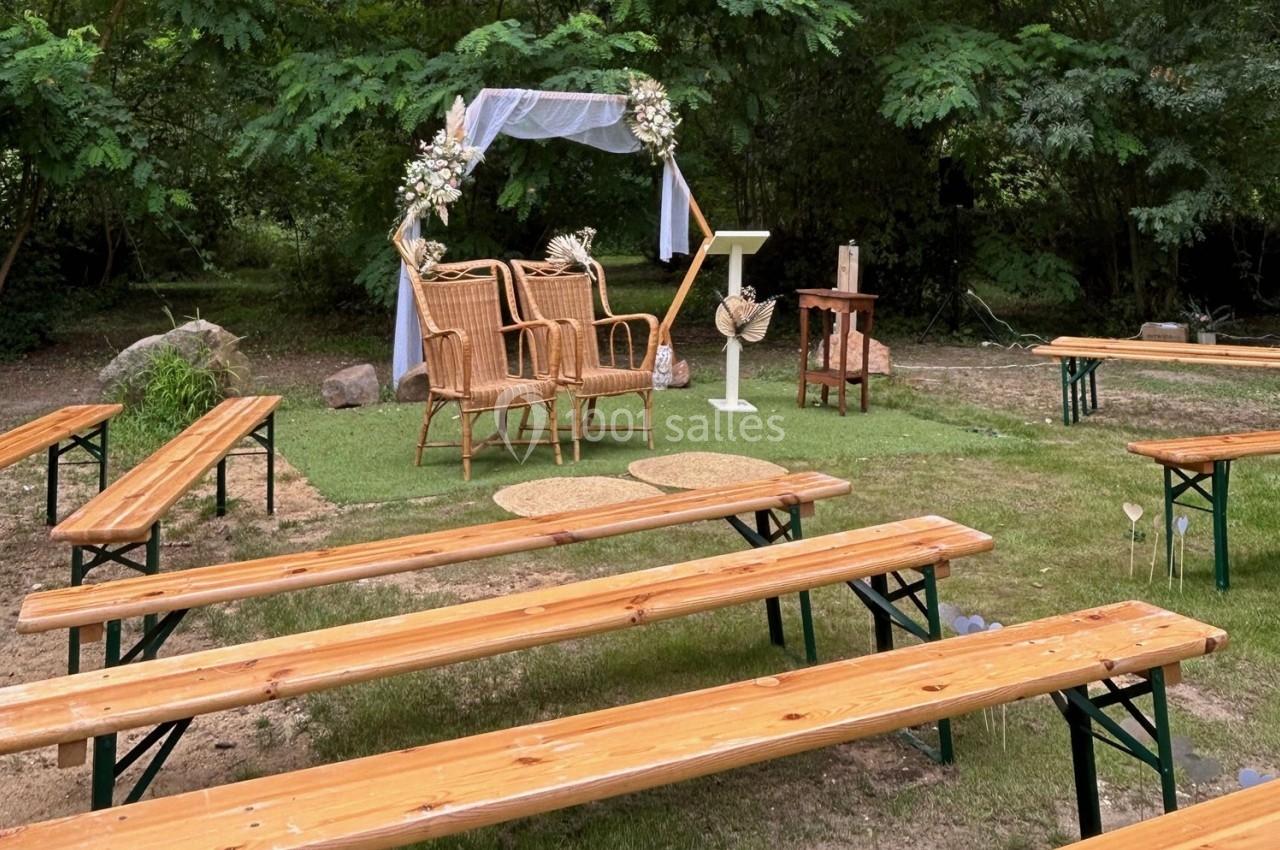 Chaises en osier et arche décorée pour une cérémonie en plein air, entourées de bancs en bois sur une pelouse.