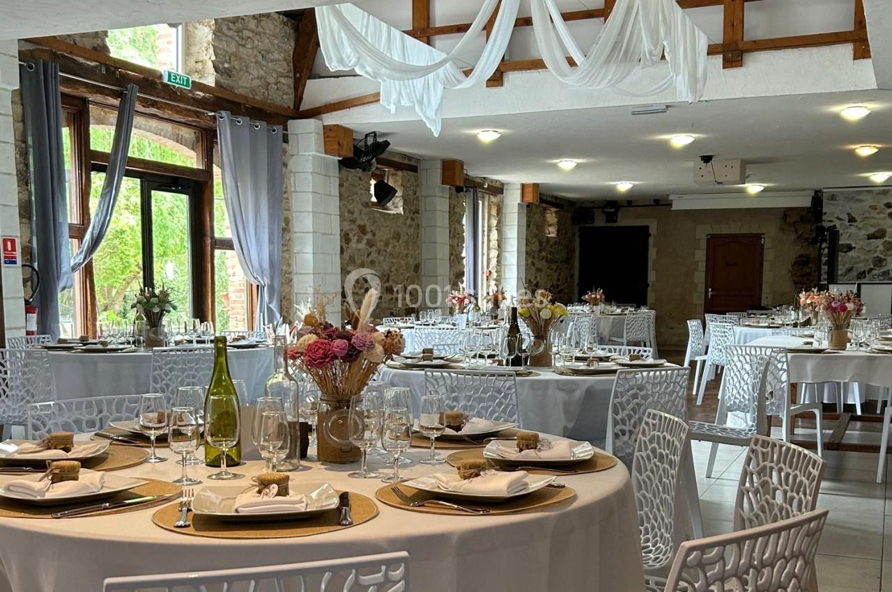 Salle de réception décorée avec des tables rondes dressées, chaises blanches et drapés suspendus au plafond.