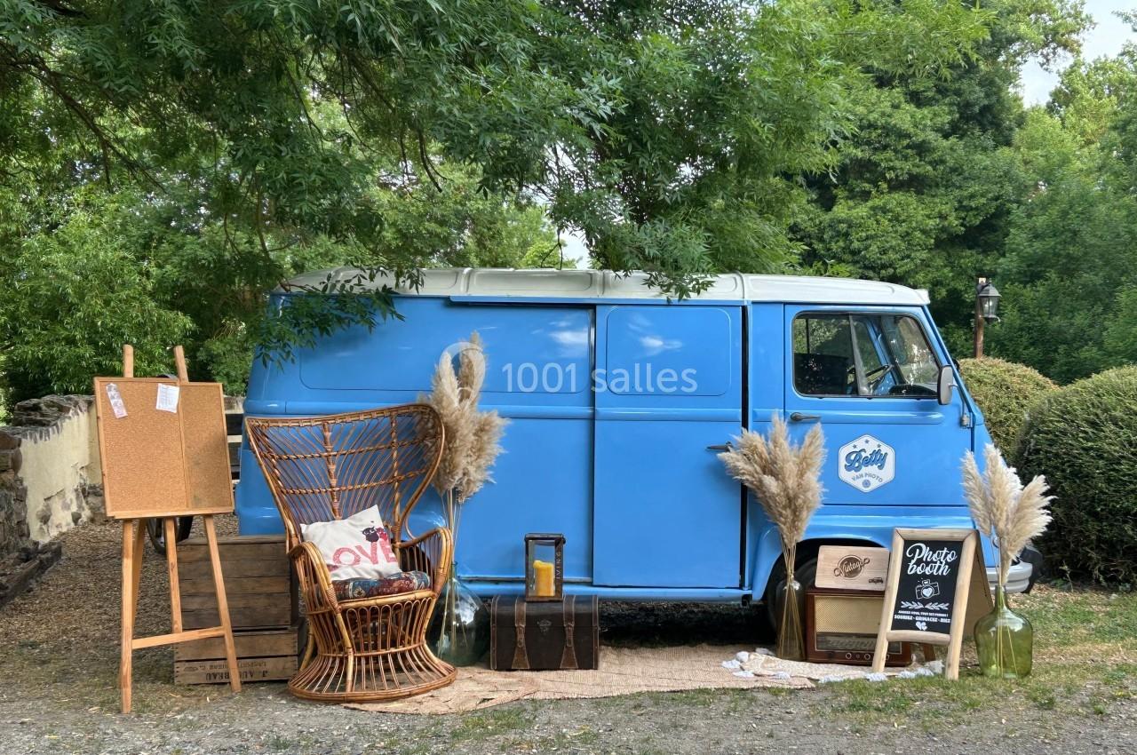 Un van bleu aménagé en photobooth, entouré de décorations vintage et végétales dans un cadre extérieur verdoyant.