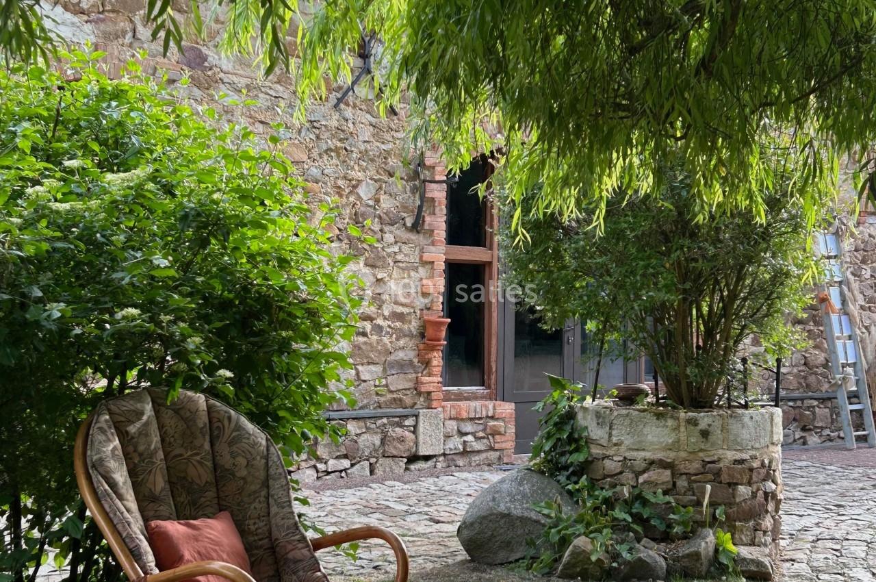 Fauteuil en rotin avec coussins sous un arbre, près d'un mur en pierre et d'une cour pavée.