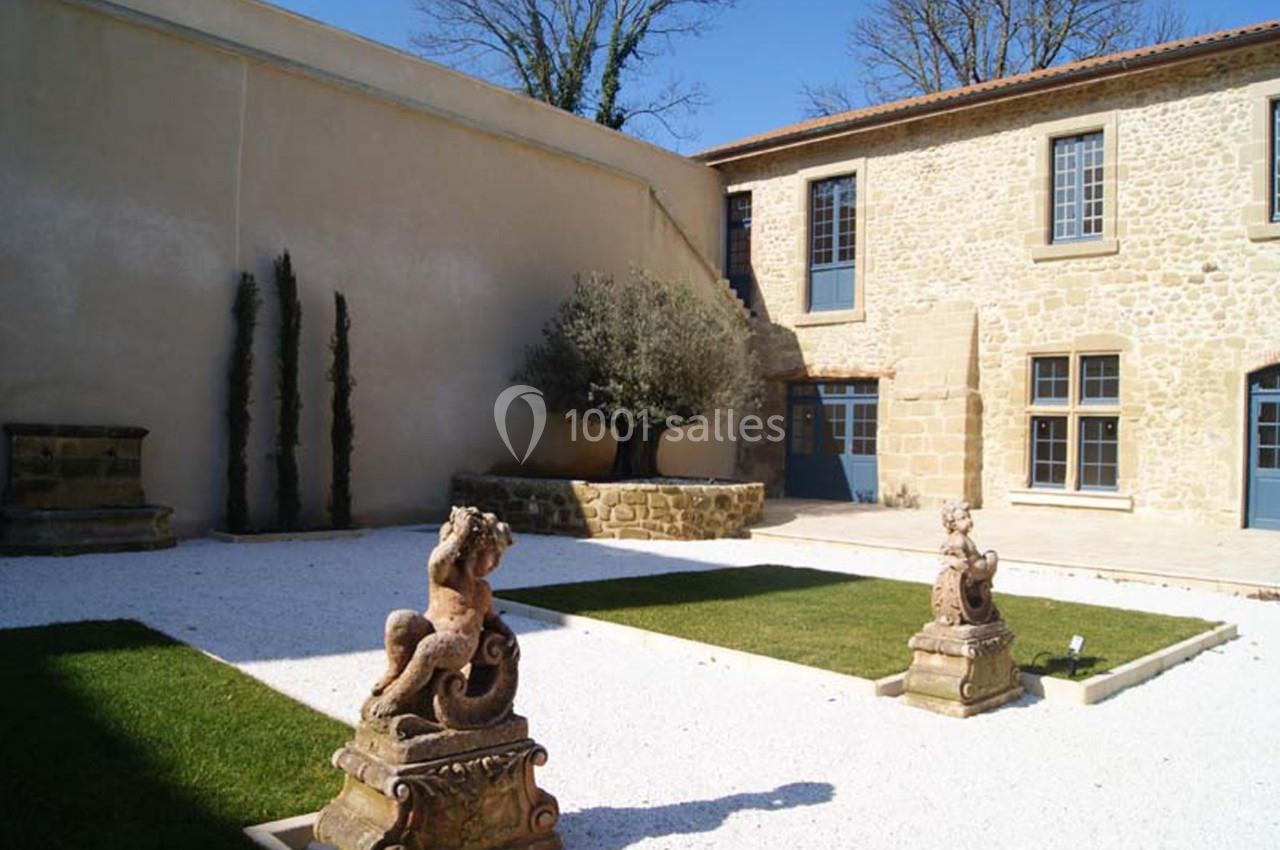 Cour intérieure avec pelouse, statues décoratives, arbre central et façade en pierre aux fenêtres bleues.