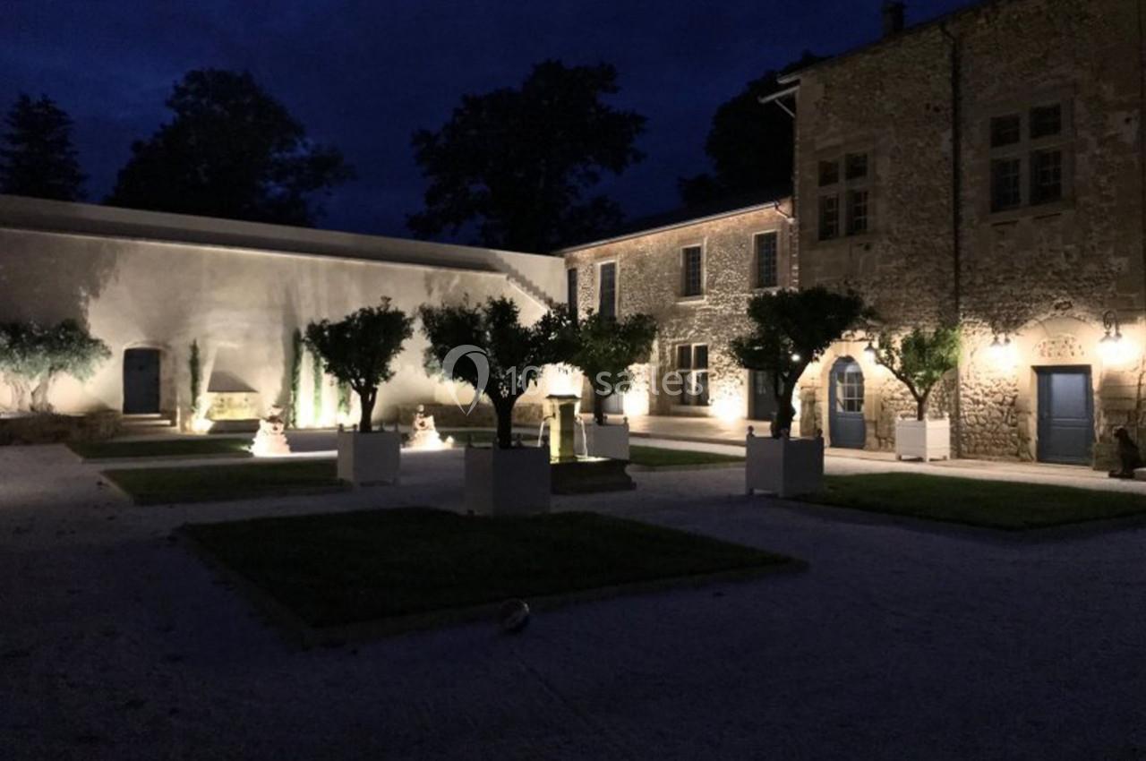 Cour intérieure d'un bâtiment en pierre éclairée la nuit, avec des arbres en pots et un éclairage chaleureux.