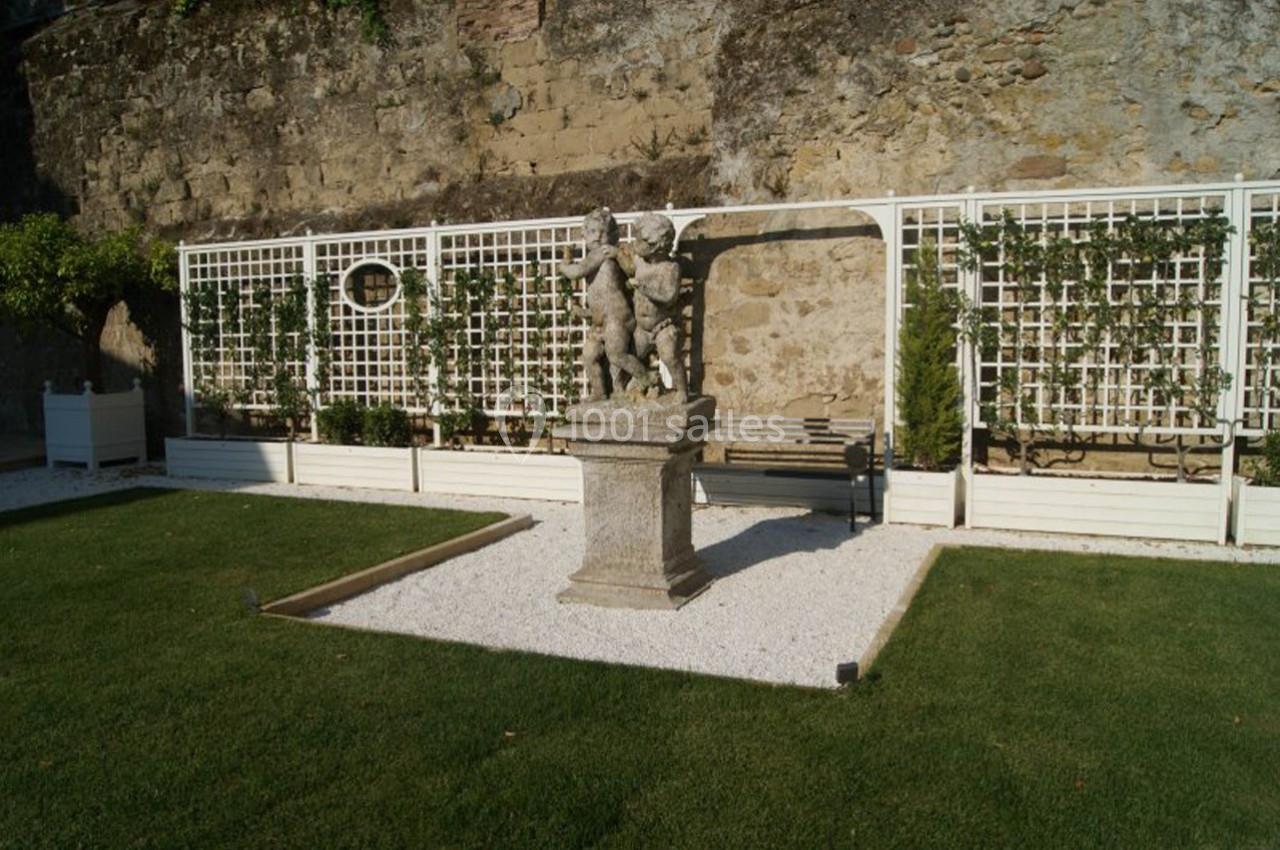 Statue en pierre sur un socle, entourée de gravier, devant une treille et un mur en pierre dans un jardin aménagé.