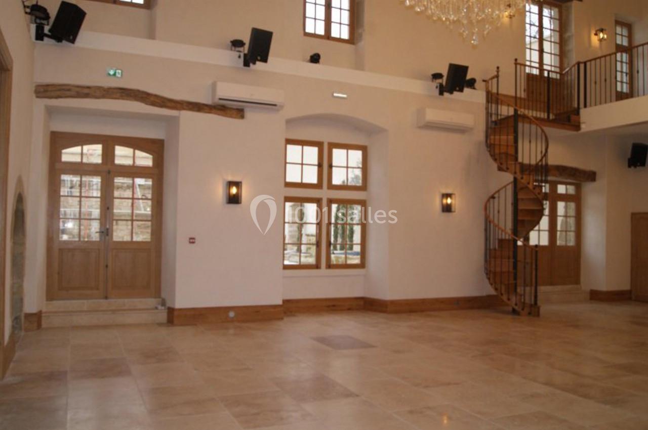 Salle spacieuse avec sol en pierre, grandes fenêtres, escalier en colimaçon en bois et murs clairs.