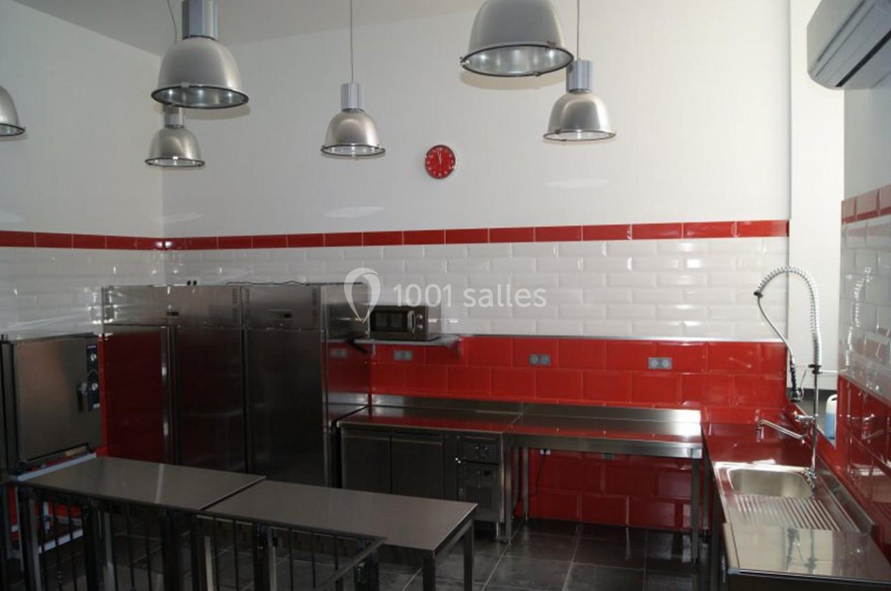 Cuisine professionnelle avec mobilier en inox, murs carrelés rouges et blancs, éclairage suspendu et horloge murale rouge.