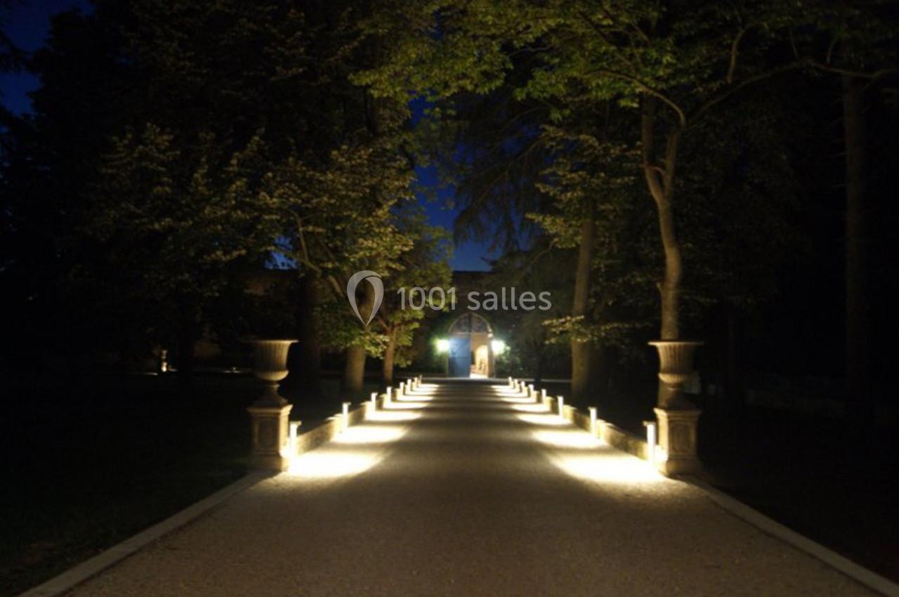 Allée illuminée bordée d'arbres et de lampadaires, menant à une porte éclairée dans un cadre nocturne.