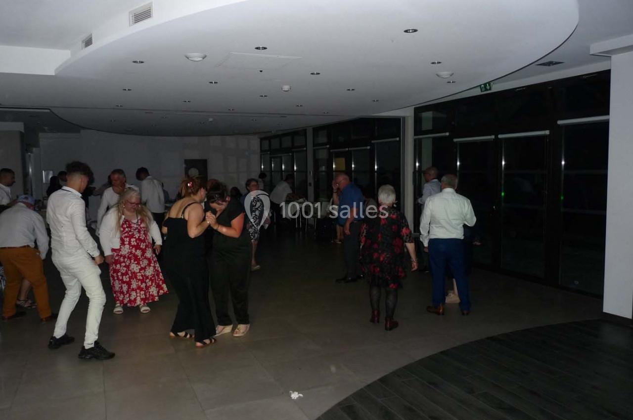 Des personnes dansent et discutent dans une salle sombre avec un sol en carrelage et des baies vitrées.