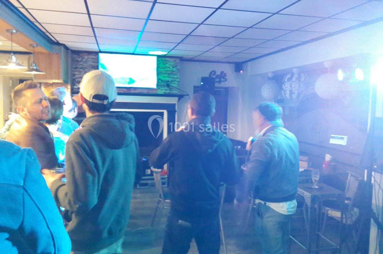 Des personnes discutent et se tiennent debout dans un bar éclairé, avec un écran allumé en arrière-plan.