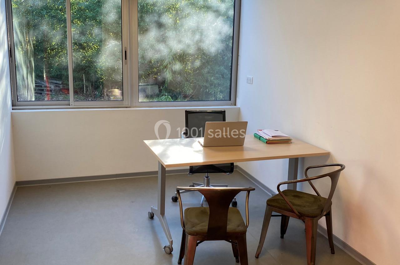 Bureau lumineux avec une table, deux chaises, un ordinateur portable et une fenêtre donnant sur un espace arboré.