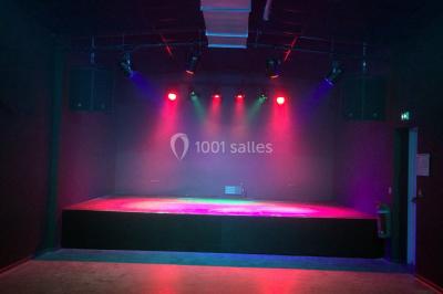 Scène vide éclairée par des projecteurs colorés dans une salle sombre avec un sol en béton.