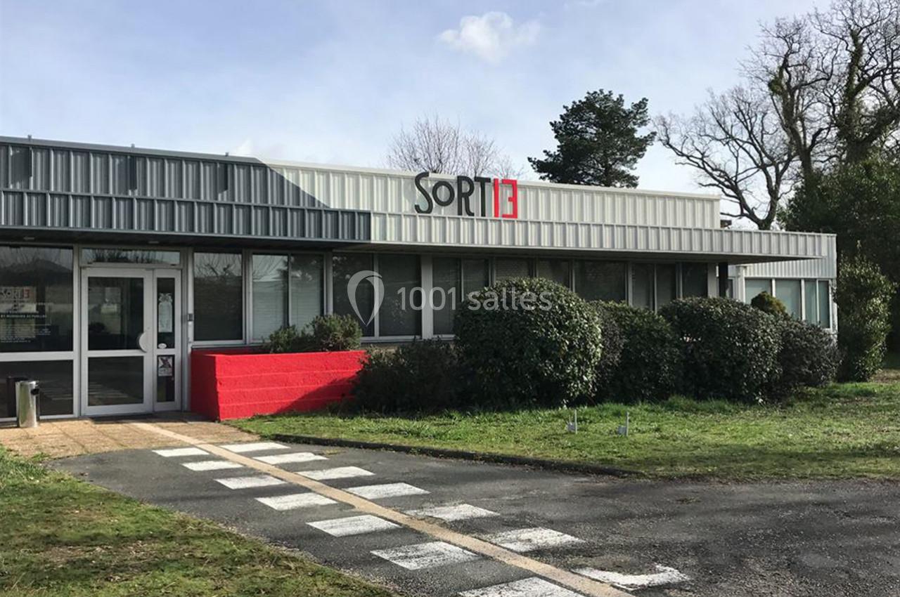 Façade d'un bâtiment moderne avec le logo ’SORTIE’, entouré de verdure et d'un passage piéton au premier plan.