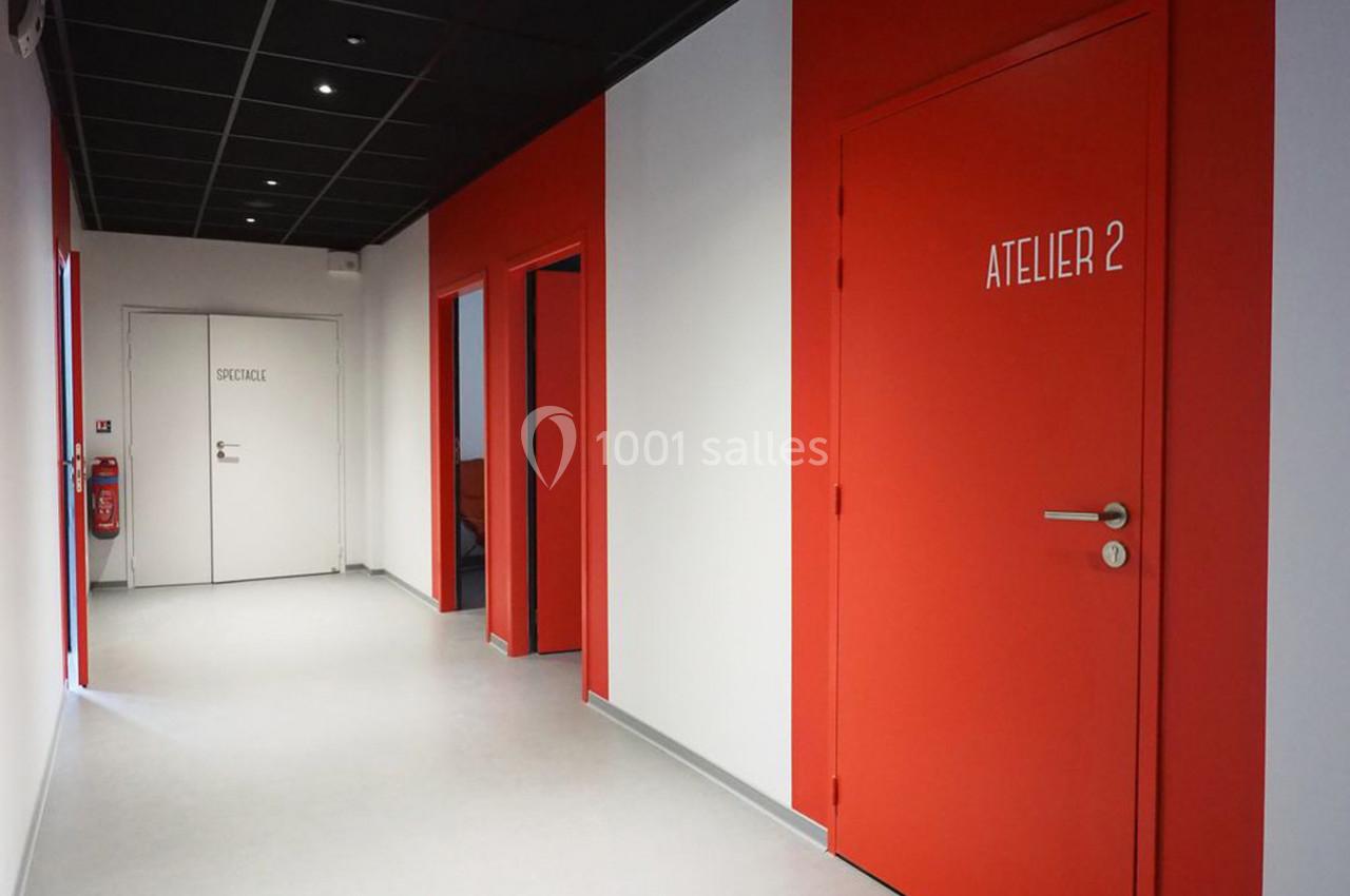 Couloir intérieur avec murs rouges et blancs, portes indiquant ’Atelier 2’ et ’Stockage’, sol clair et plafond noir.