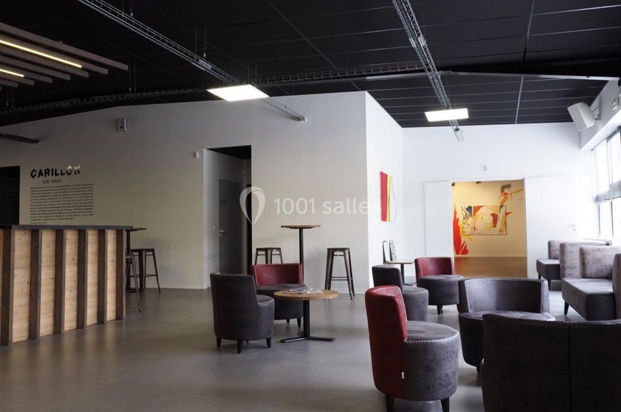 Espace lounge moderne avec fauteuils, tables basses, comptoir en bois et murs décorés, éclairé par des fenêtres.