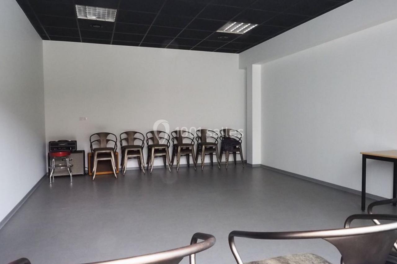 Salle vide avec des chaises alignées contre un mur blanc, sol gris et plafond noir avec éclairage fluorescent.