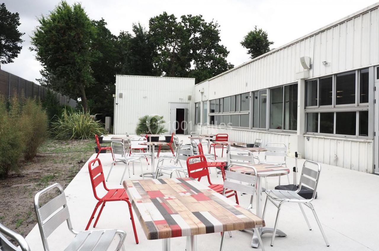 Terrasse extérieure avec tables et chaises en métal, entourée de végétation et attenante à un bâtiment blanc.