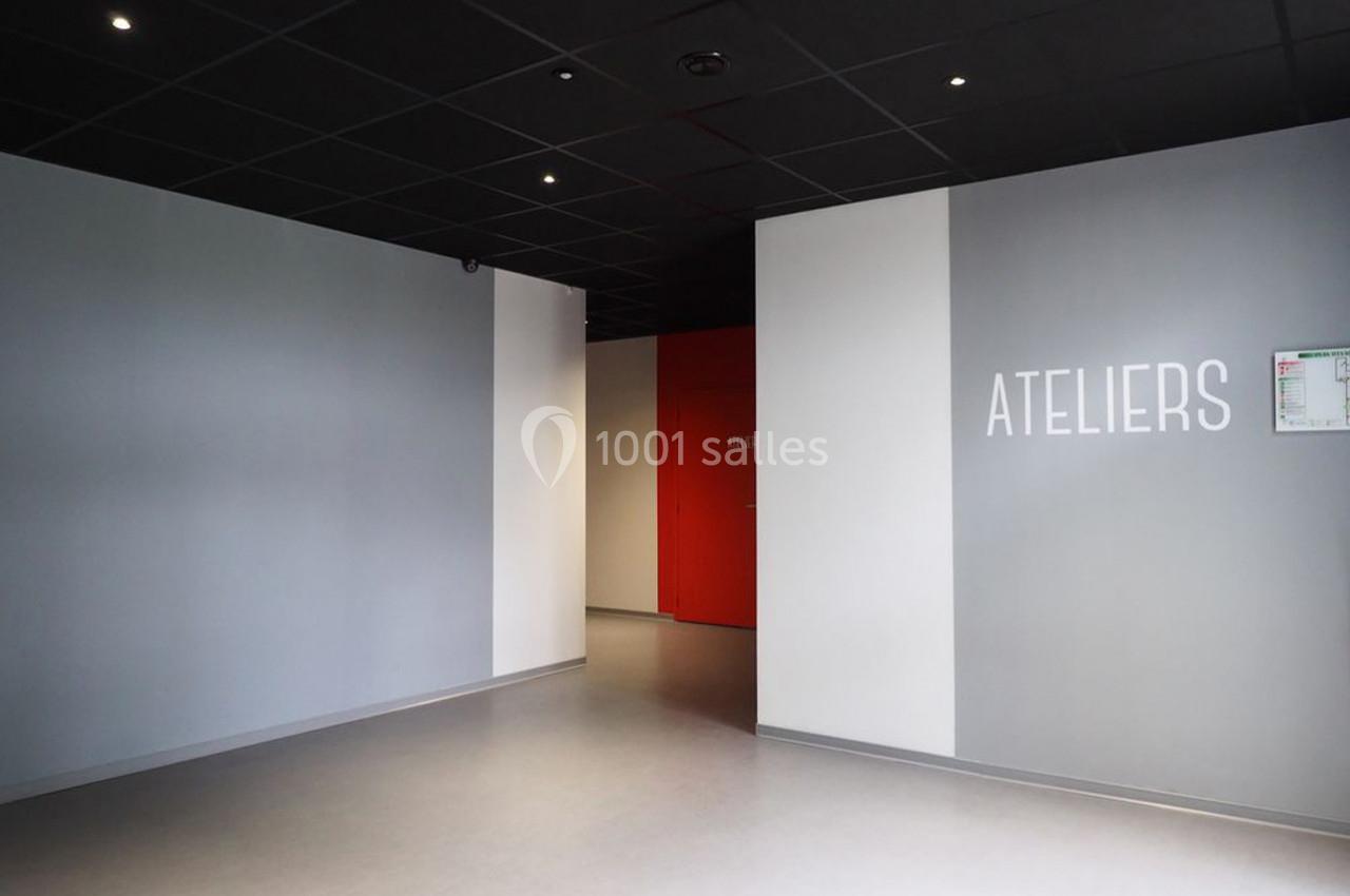 Mur gris avec l'inscription ’ATELIERS’, sol clair, plafond noir et porte rouge au fond d'un couloir.