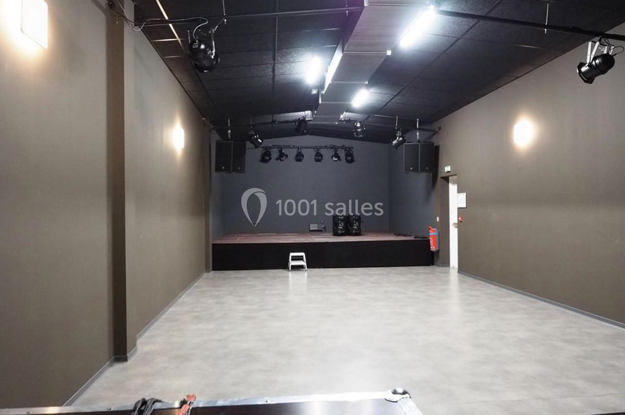 Salle vide avec sol en béton, murs sombres, éclairage au plafond et petite scène surélevée au fond.