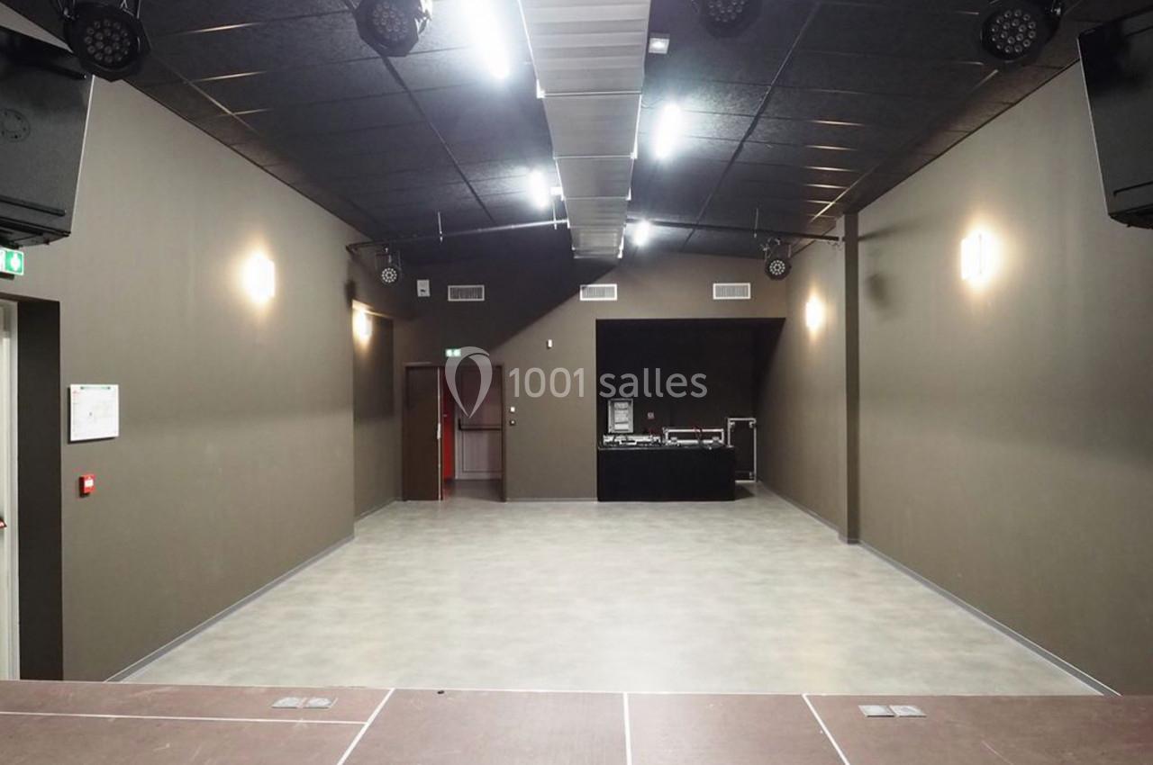 Salle vide avec sol en béton, murs sombres, éclairage au plafond et une table de mixage au fond.