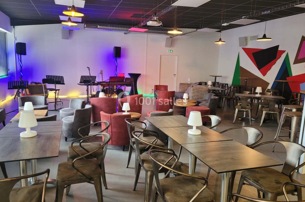 Salle moderne avec tables, chaises, fauteuils, scène équipée de micros et instruments, éclairage tamisé.