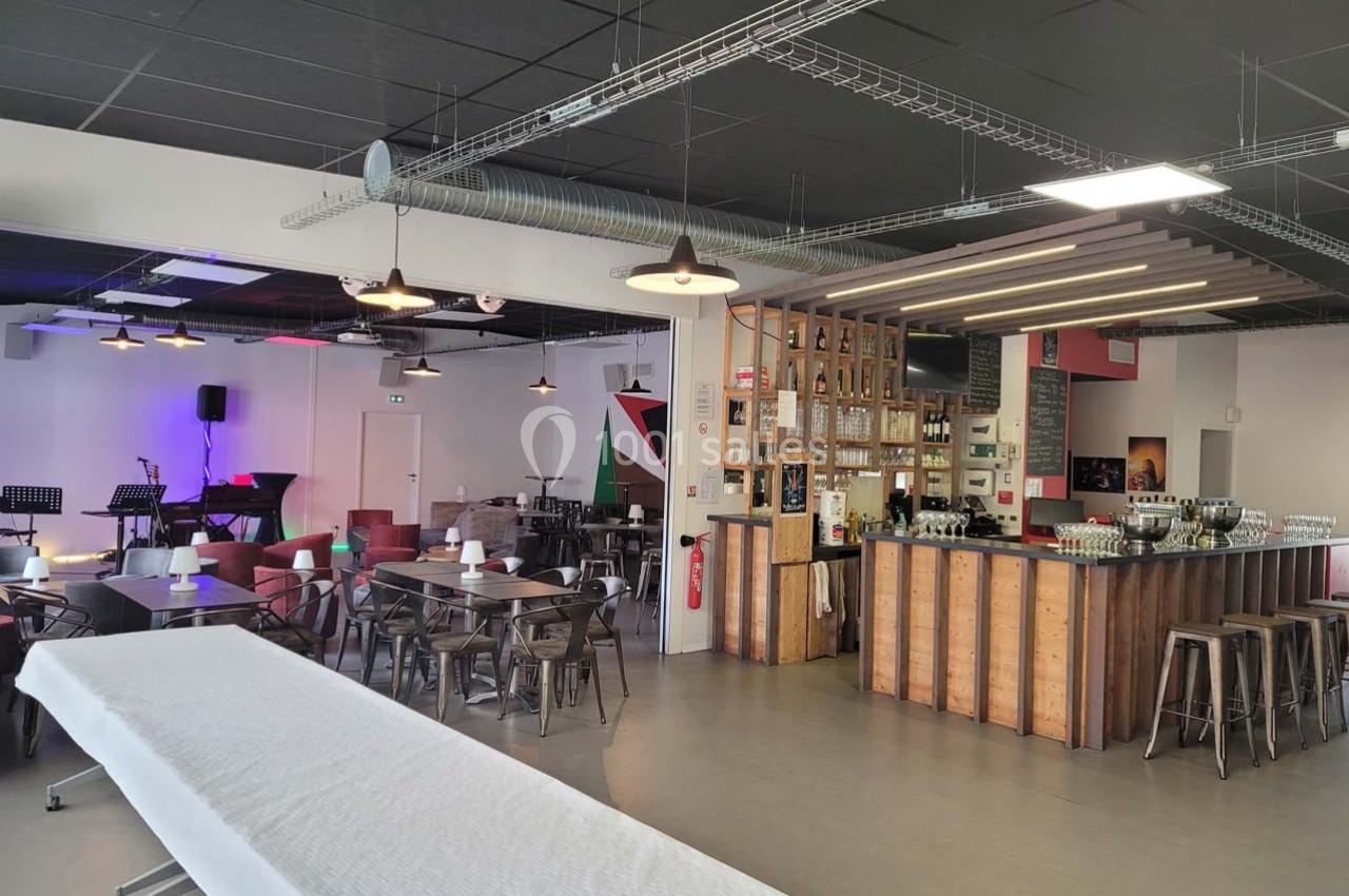 Salle moderne avec tables, chaises, bar en bois et scène équipée de micros et instruments de musique.