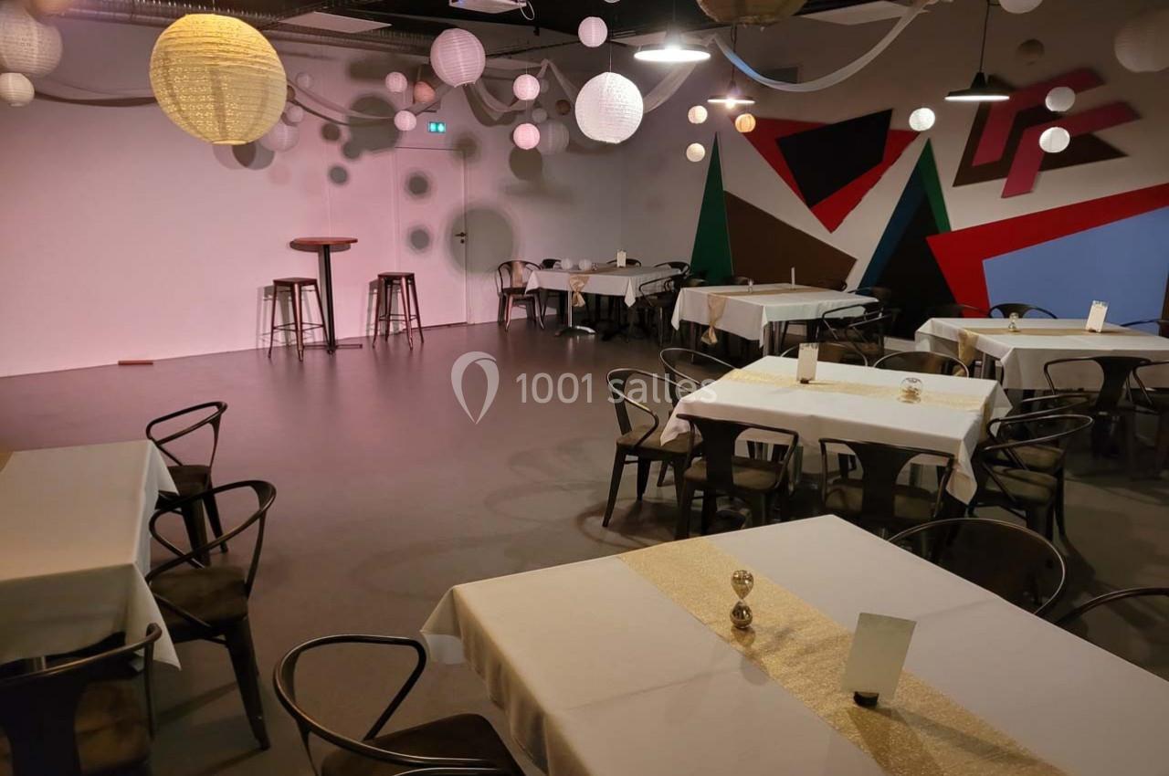 Salle décorée avec des tables dressées, des chaises noires, des lampions suspendus et un mur coloré en arrière-plan.