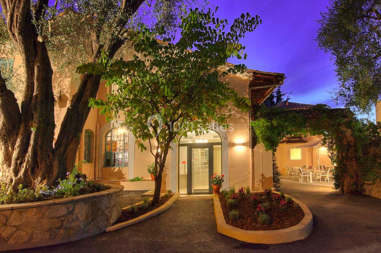 Entrée d'un bâtiment éclairé en soirée, entouré de végétation et d'arbres, avec des fenêtres et une terrasse visible.
