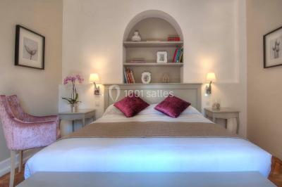 Chambre lumineuse avec un mur rose, deux fauteuils fuchsia, un lit double et des meubles blancs avec miroirs.