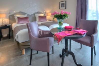 Chambre lumineuse avec un mur rose, deux fauteuils fuchsia, un lit double et des meubles blancs avec miroirs.