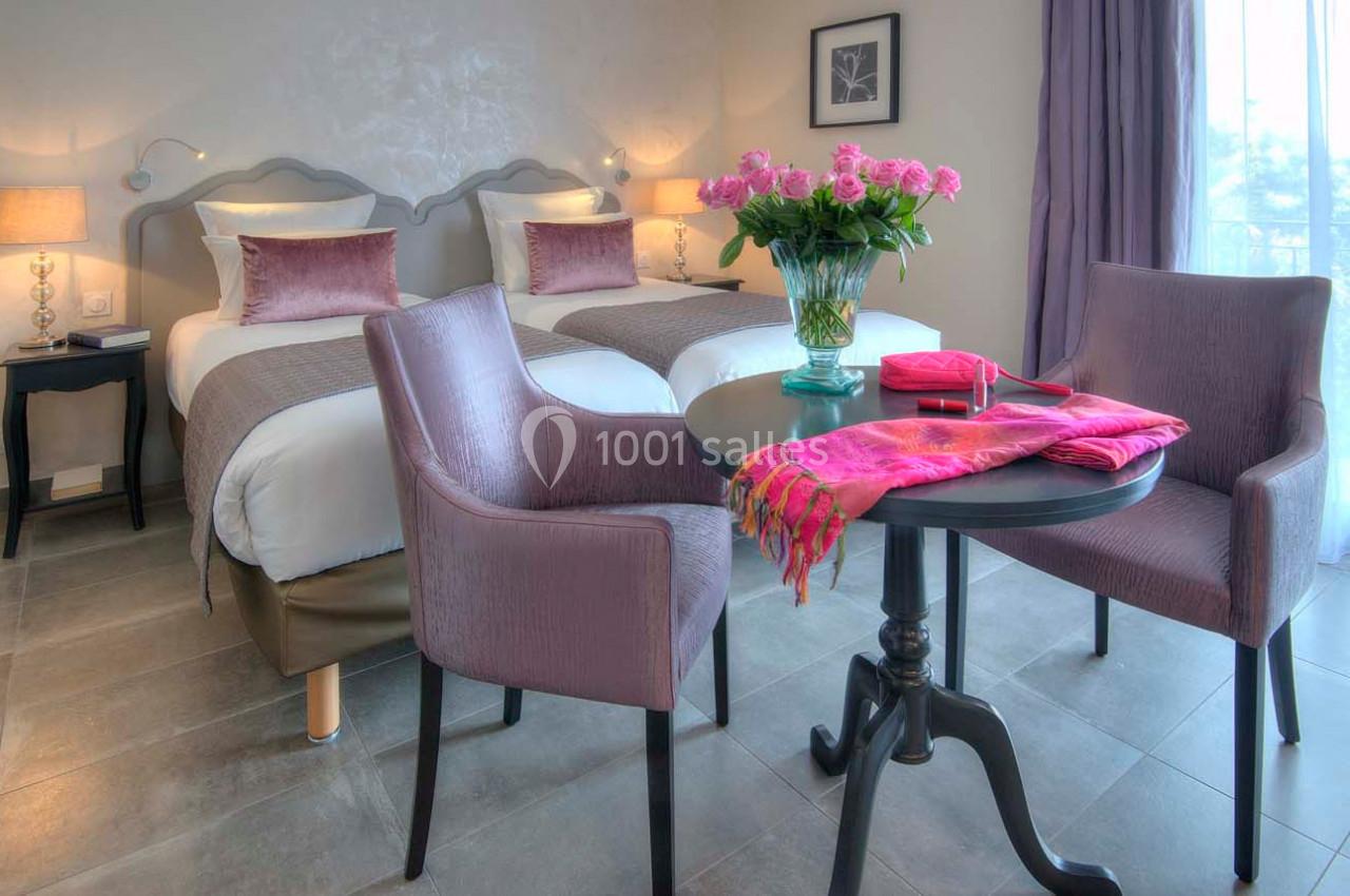 Chambre avec deux lits simples, table ronde, deux fauteuils violets et un vase de fleurs roses près d'une fenêtre.