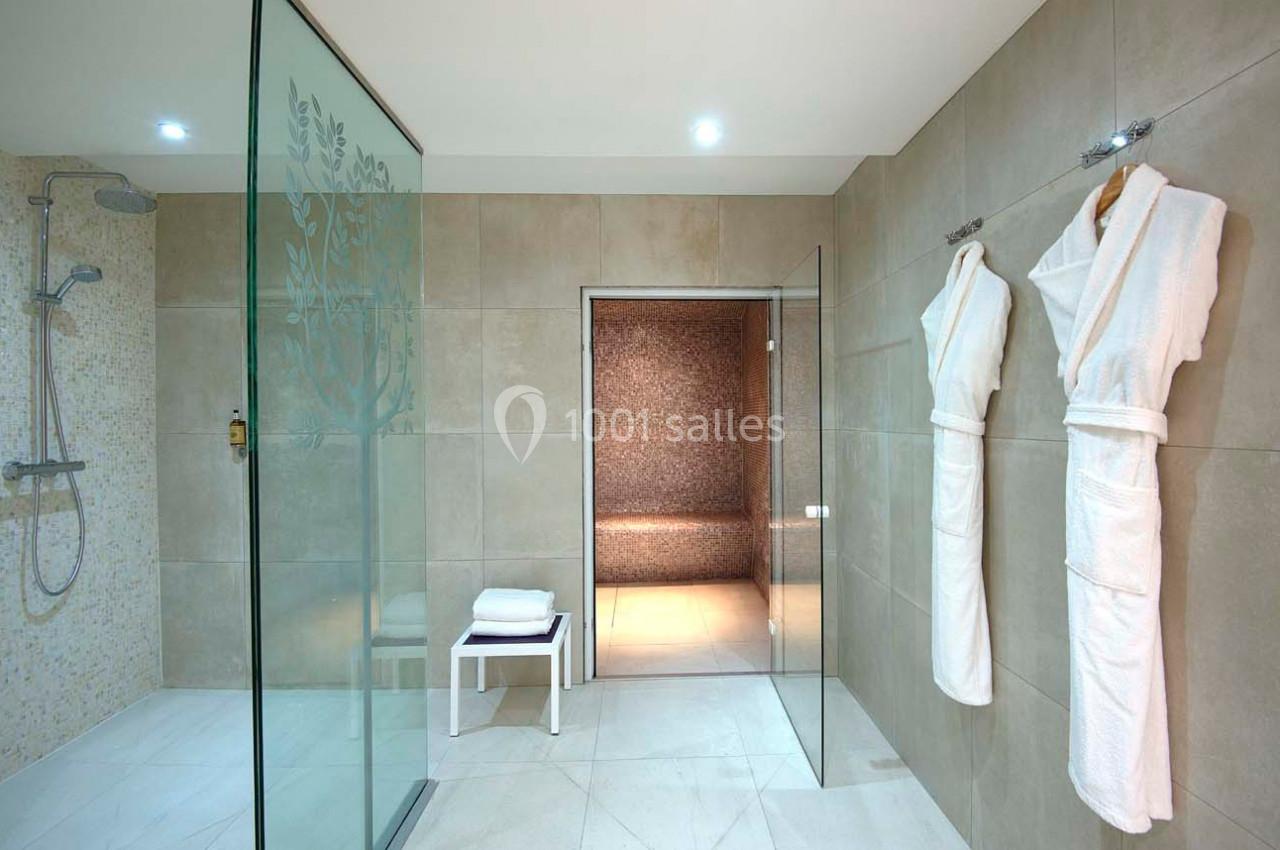 Salle de bain moderne avec douche à l'italienne, peignoirs accrochés et accès à un hammam en arrière-plan.