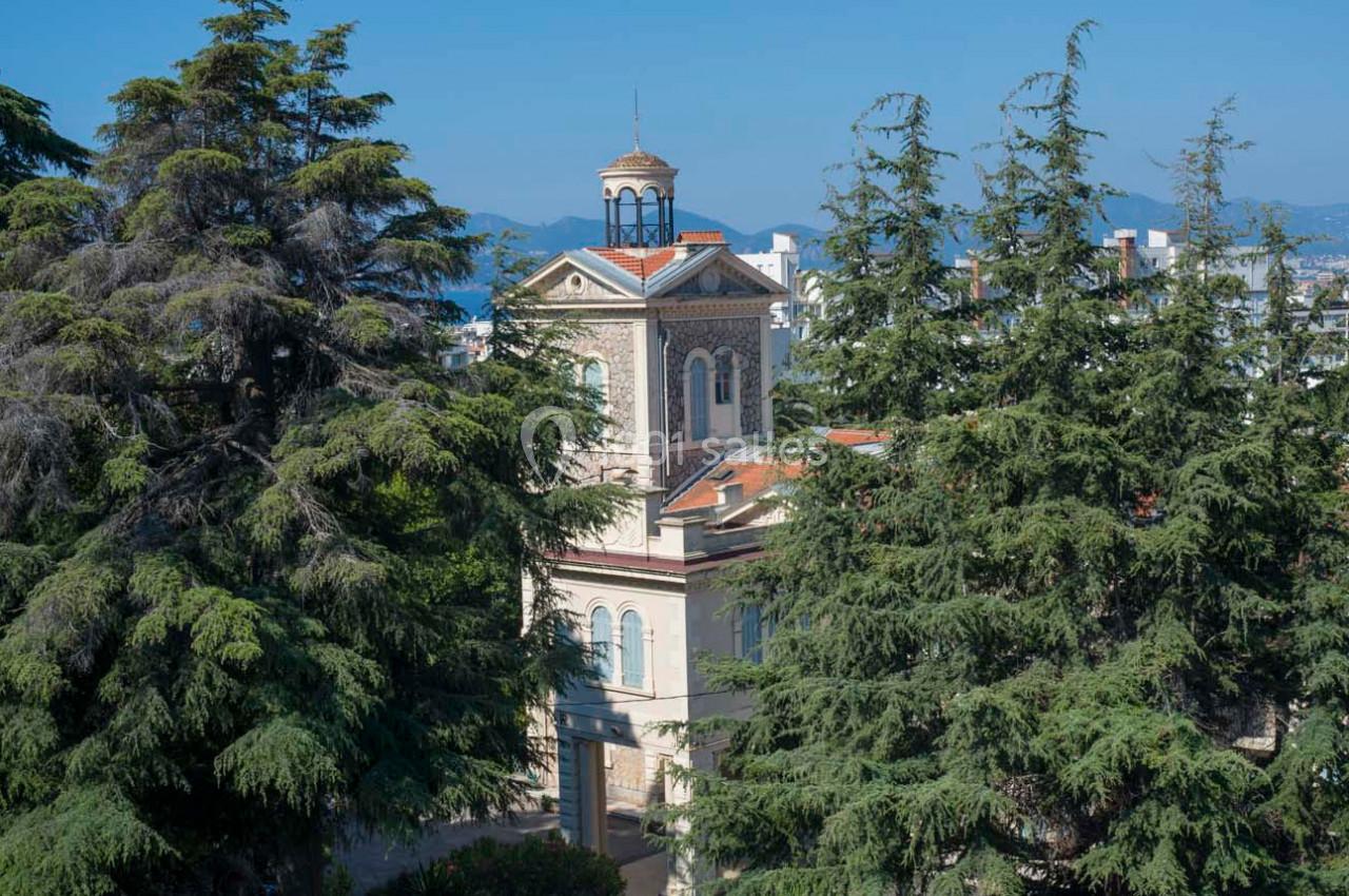 Vue d'un bâtiment ancien entouré de grands arbres, avec un ciel bleu et des collines en arrière-plan.